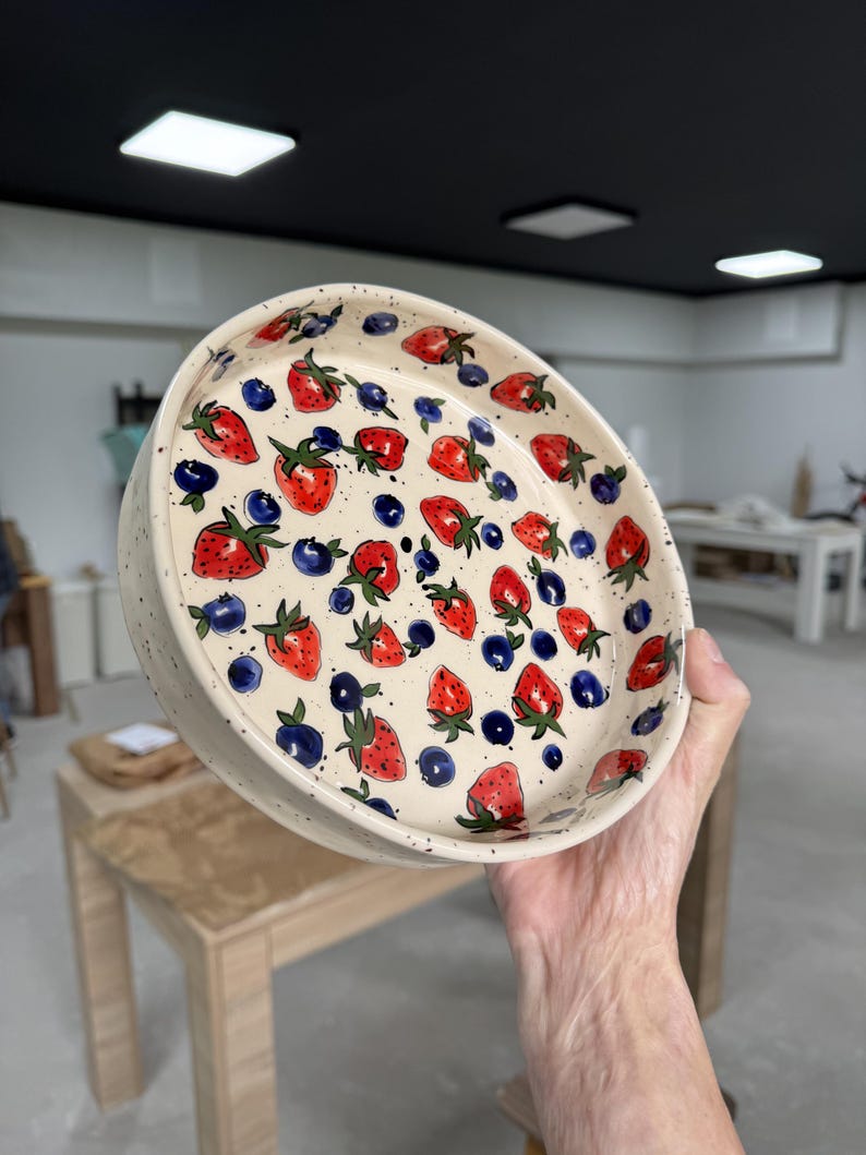 Op de afbeelding: Een ronde keramische kom met een cr&egrave;mekleurige, gespikkelde achtergrond. De kom is versierd met rode aardbeien en blauwe bosbessen. De aardbeien hebben groene steeltjes en bladeren. De kom wordt in de hand van een persoon gehouden.