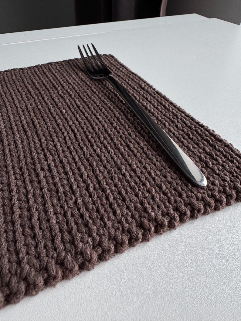 Puede incluir: Un mantel individual tejido marr&oacute;n con una superficie texturizada y un tenedor plateado encima. El mantel individual es rectangular y parece hecho a mano. El tenedor tiene un acabado pulido.