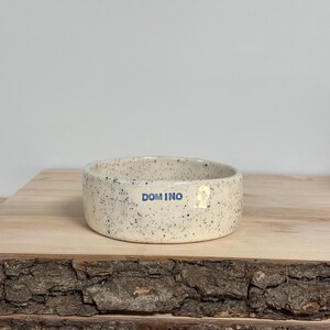 May include: A round, ceramic bowl with a speckled off-white finish and a smooth, glazed texture. The word DOMINO is printed in blue, sans-serif capital letters on the front exterior of the bowl, which has a low, straight-sided profile.