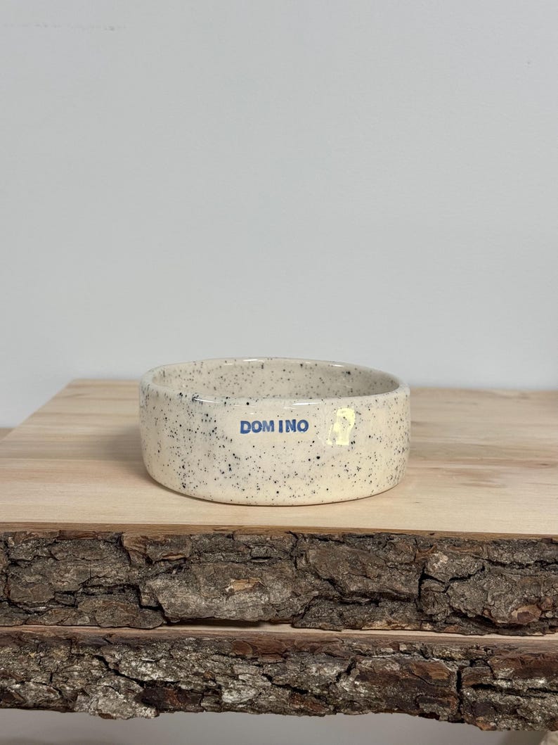 May include: A round, ceramic bowl with a speckled off-white finish and a smooth, glazed texture. The word DOMINO is printed in blue, sans-serif capital letters on the front exterior of the bowl, which has a low, straight-sided profile.