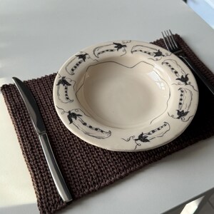 Puede incluir: Un plato de cer&aacute;mica color crema con un dise&ntilde;o de vainas de guisantes negras en el borde. Se encuentra sobre un mantel individual de punto marr&oacute;n, junto a un cuchillo de acero inoxidable y un tenedor negro. El conjunto est&aacute; sobre una superficie blanca.