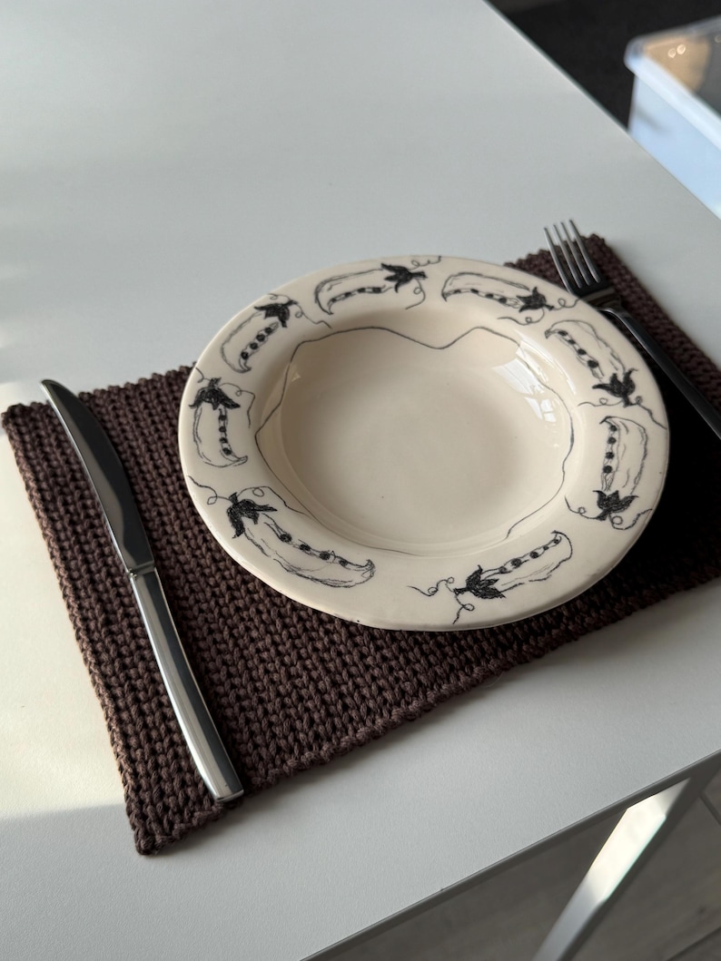 Puede incluir: Un plato de cer&aacute;mica color crema con un dise&ntilde;o de vainas de guisantes negras en el borde. Se encuentra sobre un mantel individual de punto marr&oacute;n, junto a un cuchillo de acero inoxidable y un tenedor negro. El conjunto est&aacute; sobre una superficie blanca.