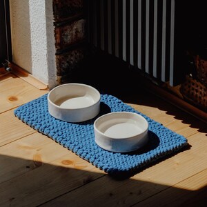 May include: Two round, white ceramic bowls are placed on a rectangular, blue, chunky knit mat. The bowls have smooth, solid-coloured surfaces and are positioned side-by-side on the textured, braided fabric base, which features a consistent weave.