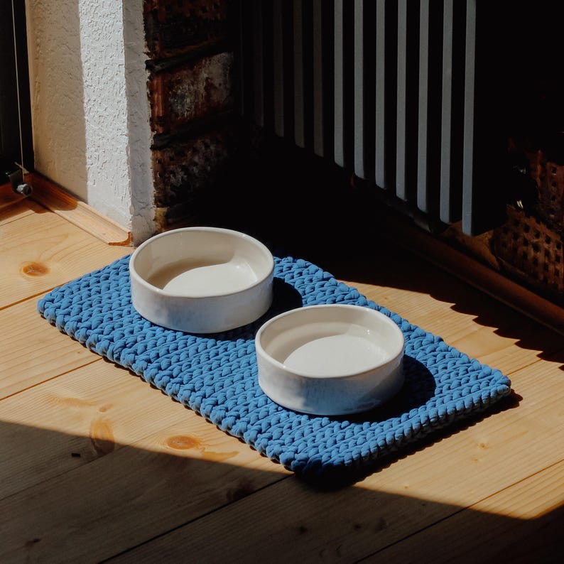 May include: Two round, white ceramic bowls are placed on a rectangular, blue, chunky knit mat. The bowls have smooth, solid-coloured surfaces and are positioned side-by-side on the textured, braided fabric base, which features a consistent weave.