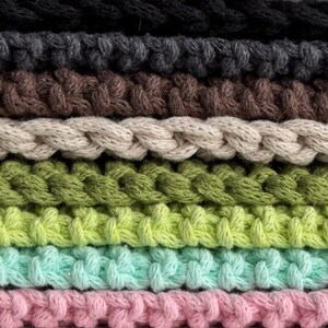 Puede incluir: Primer plano de una pila de cuerda de algod&oacute;n trenzada en varios colores. Los colores incluyen negro, gris, marr&oacute;n, beige, verde, verde claro, turquesa y rosa. La cuerda est&aacute; tejida apretadamente, creando una superficie texturizada.