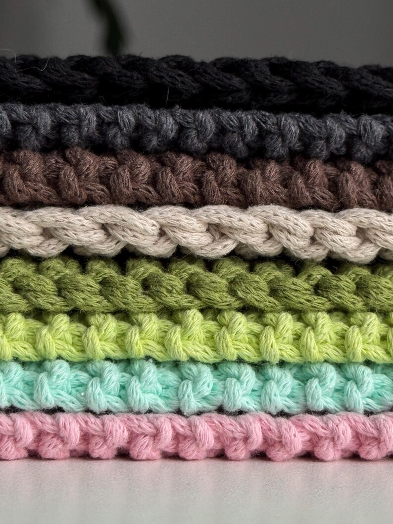 Puede incluir: Primer plano de una pila de cuerda de algod&oacute;n trenzada en varios colores. Los colores incluyen negro, gris, marr&oacute;n, beige, verde, verde claro, turquesa y rosa. La cuerda est&aacute; tejida apretadamente, creando una superficie texturizada.
