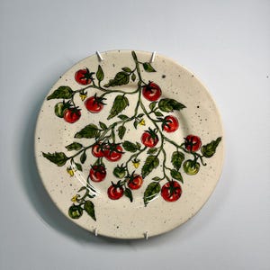 K&ouml;nnte beinhalten: Ein runder Keramikteller mit handgemaltem Design einer Tomatenpflanze. Der Teller zeigt rote und gr&uuml;ne Tomaten, gr&uuml;ne Bl&auml;tter und kleine gelbe Bl&uuml;ten auf einem gesprenkelten, cremefarbenen Hintergrund. Der Teller ist an einer wei&szlig;en Wand befestigt.