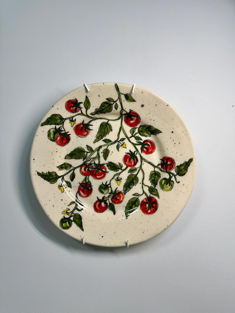 K&ouml;nnte beinhalten: Ein runder Keramikteller mit handgemaltem Design einer Tomatenpflanze. Der Teller zeigt rote und gr&uuml;ne Tomaten, gr&uuml;ne Bl&auml;tter und kleine gelbe Bl&uuml;ten auf einem gesprenkelten, cremefarbenen Hintergrund. Der Teller ist an einer wei&szlig;en Wand befestigt.