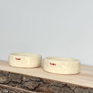 May include: Two off-white ceramic bowls with a slightly irregular, hand-formed texture. Each bowl features the name TAMI printed in small, bold red capital letters on the exterior side. The bowls have a smooth, glossy finish and low walls.