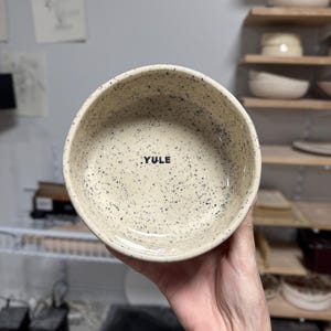 May include: A round ceramic bowl with a cream-coloured base and black speckles throughout the interior and exterior. The word YULE is printed in black, uppercase, sans-serif letters on the interior base of the bowl. The bowl has a smooth finish.