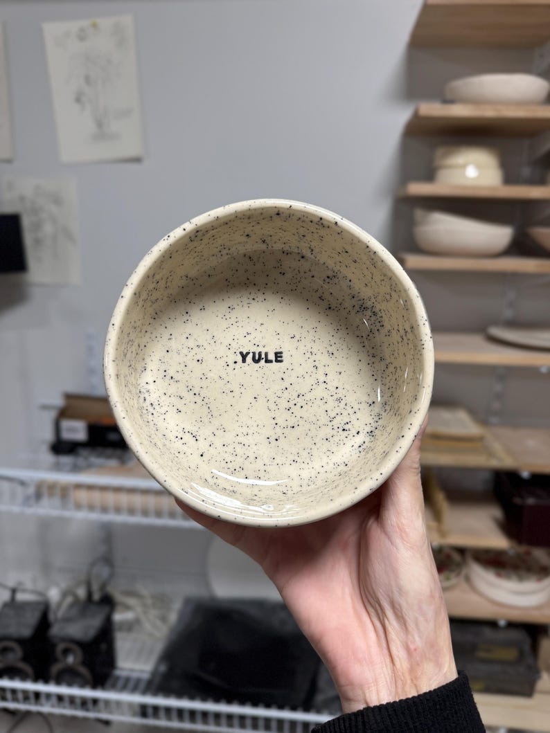 May include: A round ceramic bowl with a cream-coloured base and black speckles throughout the interior and exterior. The word YULE is printed in black, uppercase, sans-serif letters on the interior base of the bowl. The bowl has a smooth finish.