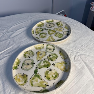 Puede incluir: Dos platos redondos de cerámica con un patrón pintado a mano de peras y manzanas verdes y amarillas. Los platos tienen una base de color blanco roto con motas, contornos oscuros y pequeños puntos negros dispersos por la superficie.