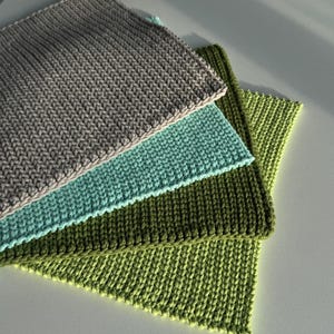 Puede incluir: Cuatro pa&ntilde;os de lavado tejidos en tonos gris, turquesa, verde oliva y verde lima est&aacute;n apilados sobre una superficie blanca. Los pa&ntilde;os tienen una textura acanalada y est&aacute;n cuidadosamente dispuestos, mostrando una est&eacute;tica artesanal.