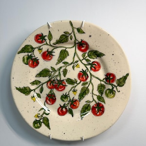 K&ouml;nnte beinhalten: Ein runder, cremefarbener Keramikteller mit einem handgemalten Design einer Tomatenranke. Die Ranke hat gr&uuml;ne Bl&auml;tter, rote und gr&uuml;ne Tomaten und kleine gelbe Bl&uuml;ten. Der Teller hat eine gesprenkelte Textur und ist an einer wei&szlig;en Wand befestigt.