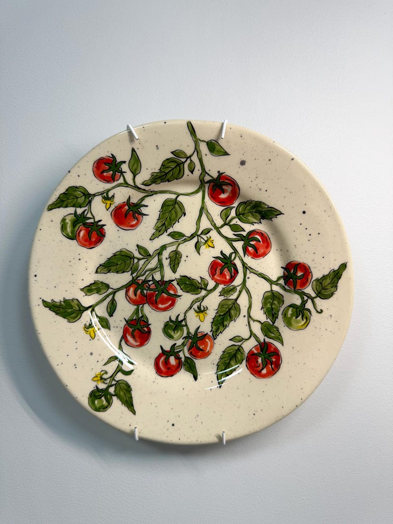 K&ouml;nnte beinhalten: Ein runder, cremefarbener Keramikteller mit einem handgemalten Design einer Tomatenranke. Die Ranke hat gr&uuml;ne Bl&auml;tter, rote und gr&uuml;ne Tomaten und kleine gelbe Bl&uuml;ten. Der Teller hat eine gesprenkelte Textur und ist an einer wei&szlig;en Wand befestigt.