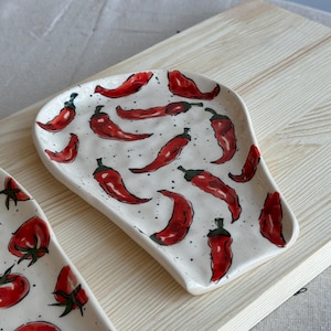 May include: White ceramic spoon rest with a hand-painted red chili pepper design. The peppers are scattered across the surface, with black speckles adding detail. A second spoon rest with a tomato design is also visible.