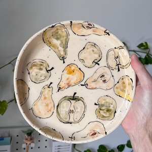 Op de afbeelding: Een ronde keramische plaat met een crèmekleurige achtergrond, versierd met handgeschilderde peren in verschillende stadia van rijpheid. De peren zijn in tinten groen, geel en bruin, omlijnd in zwart. De plaat heeft een gespikkeld ontwerp.