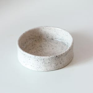 May include: A small, round ceramic bowl with a white base and a speckled pattern featuring numerous tiny black dots distributed across the interior and exterior surfaces. The bowl has a smooth, glossy finish and straight, vertical sides.
