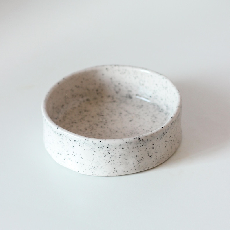 May include: A small, round ceramic bowl with a white base and a speckled pattern featuring numerous tiny black dots distributed across the interior and exterior surfaces. The bowl has a smooth, glossy finish and straight, vertical sides.