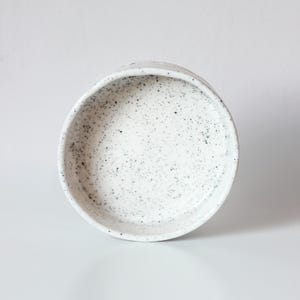 May include: A round, shallow ceramic bowl with a white base and a speckled pattern of small black dots across the interior surface. The bowl features a slightly uneven, organic rim and a smooth, matte finish throughout the entire piece.