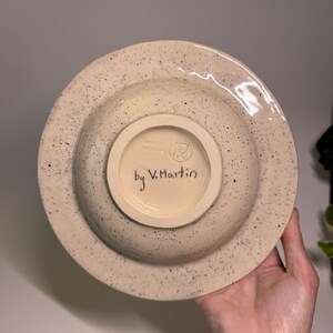 Puede incluir: Un cuenco de cer&aacute;mica beige moteado con un borde ancho. La parte inferior presenta un c&iacute;rculo m&aacute;s peque&ntilde;o y empotrado con el texto "by V. Martin" escrito en escritura negra. El cuenco tiene un patr&oacute;n moteado de puntos negros.