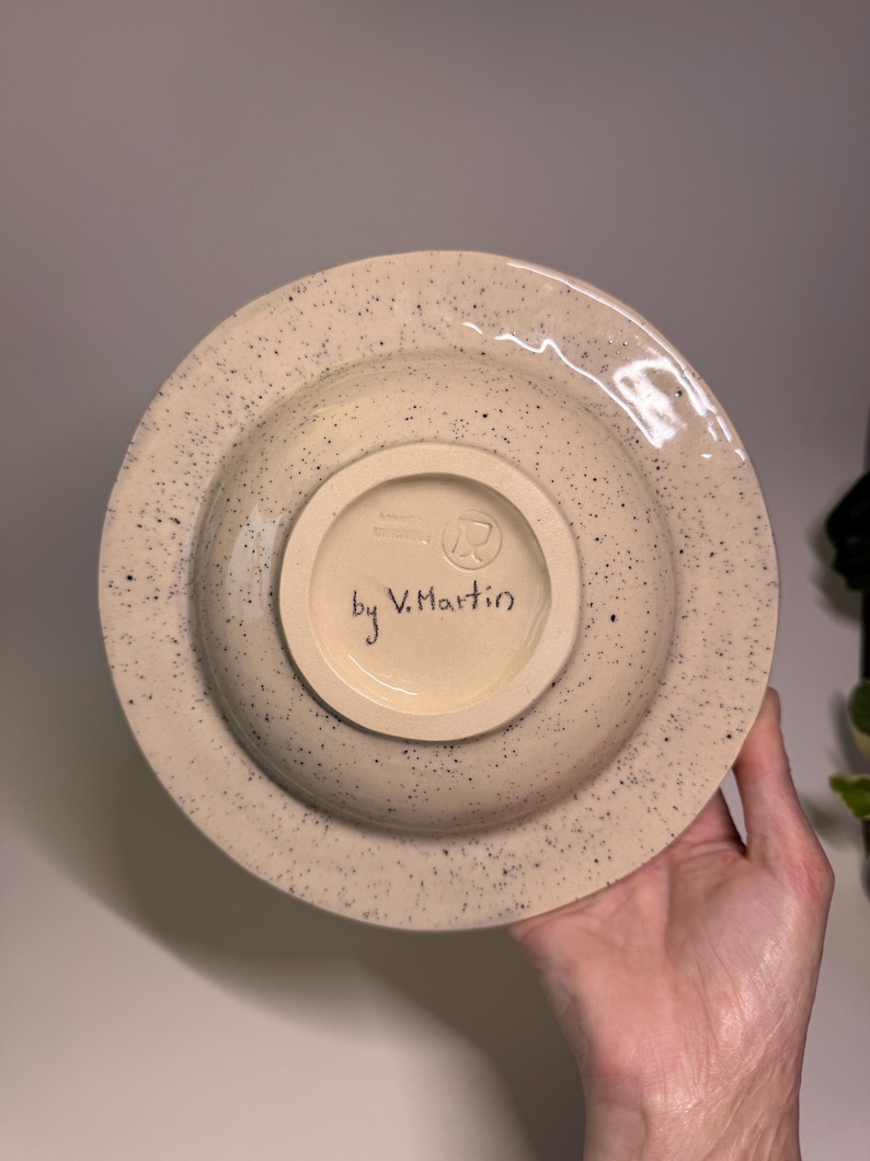 Puede incluir: Un cuenco de cer&aacute;mica beige moteado con un borde ancho. La parte inferior presenta un c&iacute;rculo m&aacute;s peque&ntilde;o y empotrado con el texto "by V. Martin" escrito en escritura negra. El cuenco tiene un patr&oacute;n moteado de puntos negros.