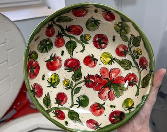 May include: A ceramic bowl with a hand-painted tomato design. The bowl features a cream-colored interior with red and green tomatoes, green leaves, and a green rim. The tomatoes vary in size and color, with some yellow and green.