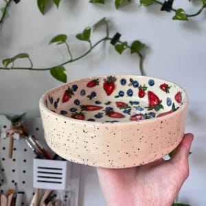 Op de afbeelding: Een cr&egrave;mekleurige keramische kom met zwarte spikkels, versierd met rode aardbeien en blauwe bosbessen. De kom wordt in een hand gehouden, met een witte muur en groene wijnranken op de achtergrond. De diameter van de kom is ongeveer 15 cm.