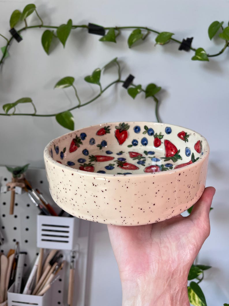 Op de afbeelding: Een cr&egrave;mekleurige keramische kom met zwarte spikkels, versierd met rode aardbeien en blauwe bosbessen. De kom wordt in een hand gehouden, met een witte muur en groene wijnranken op de achtergrond. De diameter van de kom is ongeveer 15 cm.