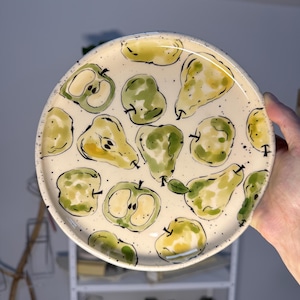 May include: A round ceramic plate with a cream-colored base featuring a hand-painted pattern of green and yellow pears and apples. The design includes black ink outlines and small black speckles scattered across the surface of the plate.