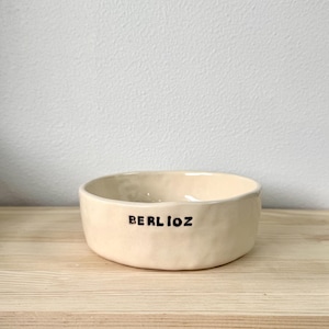 May include: A cream-coloured ceramic bowl with a slightly uneven, hand-formed texture. The exterior features the word BERLIOZ printed in bold, black, uppercase letters. The bowl has a wide, open top and a low profile, showcasing a simple design.