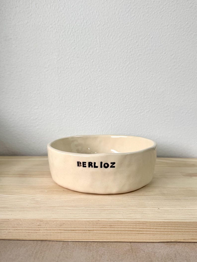 May include: A cream-coloured ceramic bowl with a slightly uneven, hand-formed texture. The exterior features the word BERLIOZ printed in bold, black, uppercase letters. The bowl has a wide, open top and a low profile, showcasing a simple design.
