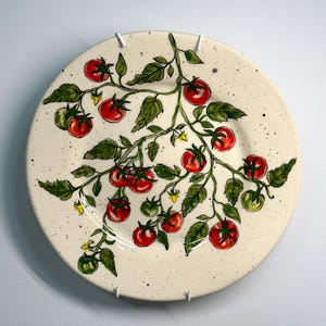 K&ouml;nnte beinhalten: Ein runder, cremefarbener Keramikteller mit handgemaltem Design einer Tomatenranke. Die Ranke hat rote und gr&uuml;ne Tomaten, gr&uuml;ne Bl&auml;tter und kleine gelbe Bl&uuml;ten. Der Teller hat eine gesprenkelte Textur und ist an einer wei&szlig;en Wand befestigt.