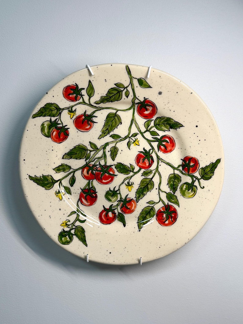 K&ouml;nnte beinhalten: Ein runder, cremefarbener Keramikteller mit handgemaltem Design einer Tomatenranke. Die Ranke hat rote und gr&uuml;ne Tomaten, gr&uuml;ne Bl&auml;tter und kleine gelbe Bl&uuml;ten. Der Teller hat eine gesprenkelte Textur und ist an einer wei&szlig;en Wand befestigt.