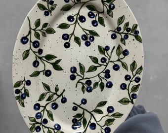 May include: Oval ceramic serving platter with a hand-painted blueberry design. The white platter features dark blue blueberries and green leaves on delicate branches. The design is speckled with small black dots.