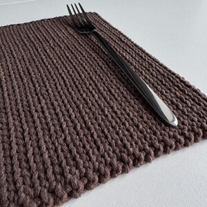 Puede incluir: Un mantel individual tejido marr&oacute;n con una superficie texturizada y un tenedor plateado encima. El mantel individual es rectangular y parece hecho a mano. El tenedor est&aacute; colocado en diagonal sobre la alfombra, lo que sugiere un entorno de comedor.