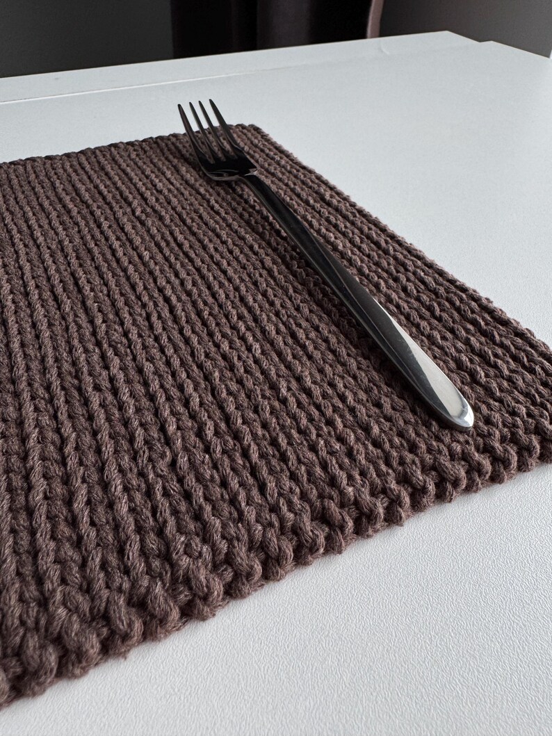 Puede incluir: Un mantel individual tejido marr&oacute;n con una superficie texturizada y un tenedor plateado encima. El mantel individual es rectangular y parece hecho a mano. El tenedor est&aacute; colocado en diagonal sobre la alfombra, lo que sugiere un entorno de comedor.