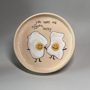 May include: A round, peach-colored ceramic plate with a hand-drawn illustration of two fried eggs holding hands. The eggs have smiling faces and the text "you make me eggstra happy" is written above them.