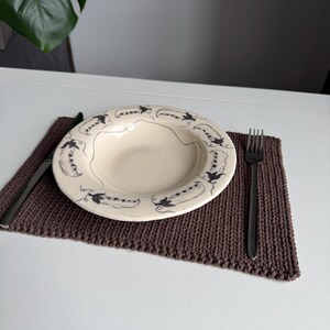 Puede incluir: Un plato de cer&aacute;mica color crema con un dise&ntilde;o de vaina de guisantes negro descansa sobre un mantel individual de punto marr&oacute;n. Un tenedor y un cuchillo se colocan a cada lado del plato. El entorno est&aacute; sobre una mesa blanca.