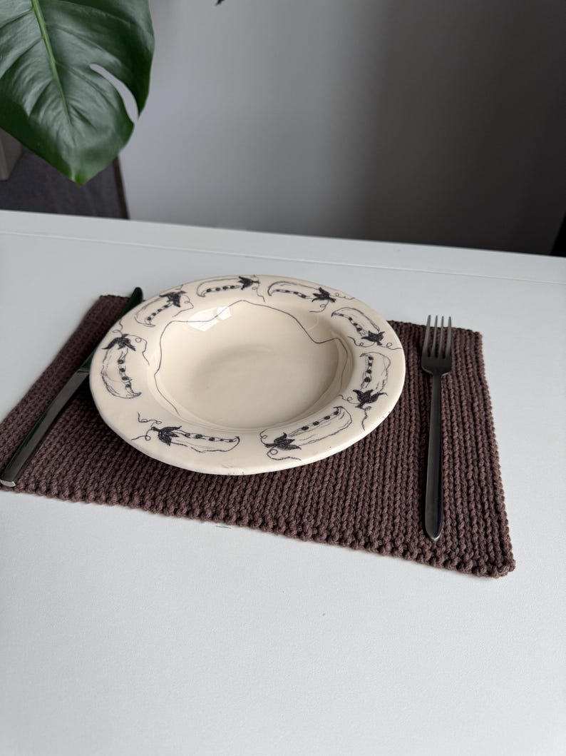 Puede incluir: Un plato de cer&aacute;mica color crema con un dise&ntilde;o de vaina de guisantes negro descansa sobre un mantel individual de punto marr&oacute;n. Un tenedor y un cuchillo se colocan a cada lado del plato. El entorno est&aacute; sobre una mesa blanca.
