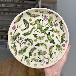 Puede incluir: Un plato redondo de cerámica con base color crema que presenta un patrón pintado a mano de vainas de guisantes verdes, enredaderas y pequeñas flores moradas. El diseño incluye guisantes verdes dispersos y sutiles motas oscuras.