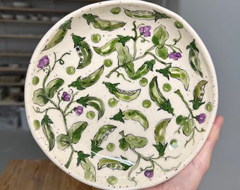 Puede incluir: Un plato redondo de cerámica con base color crema que presenta un patrón pintado a mano de vainas de guisantes verdes, enredaderas y pequeñas flores moradas. El diseño incluye guisantes verdes dispersos y sutiles motas oscuras.