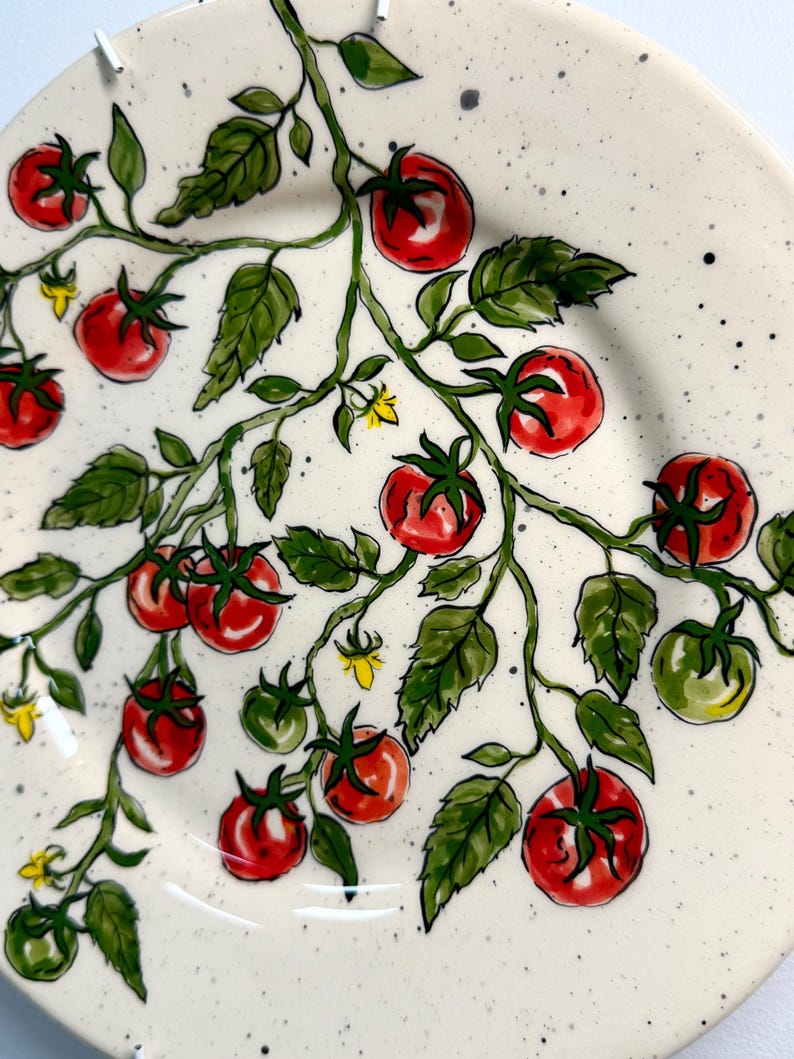 K&ouml;nnte beinhalten: Ein wei&szlig;er Keramikteller mit handgemaltem Tomatenranken-Design. Der Teller zeigt rote und gr&uuml;ne Tomaten, gr&uuml;ne Bl&auml;tter und kleine gelbe Bl&uuml;ten. Der Hintergrund hat ein gesprenkeltes Muster.