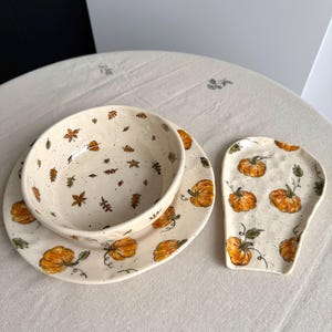 Puede incluir: Un juego de vajilla de cerámica que incluye un cuenco y un plato con esmalte crema moteado, decorado con calabazas naranjas y hojas marrones pintadas a mano. Junto al plato se encuentra un reposacucharas a juego.