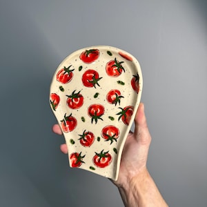 Puede incluir: Apoyacucharas de cerámica color crema con un patrón repetido de tomates rojos y hojas verdes. El apoyacucharas tiene una forma única con una parte superior ancha y curvada y un mango estrecho. Los tomates están pintados a mano con detalles en negro.