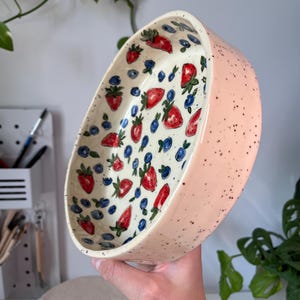 Op de afbeelding: Een ovale keramische kom met een lichtroze buitenkant, bespikkeld met zwarte stippen. De binnenkant heeft een witte achtergrond met een patroon van rode aardbeien en blauwe bosbessen. De kom wordt in een hand gehouden.