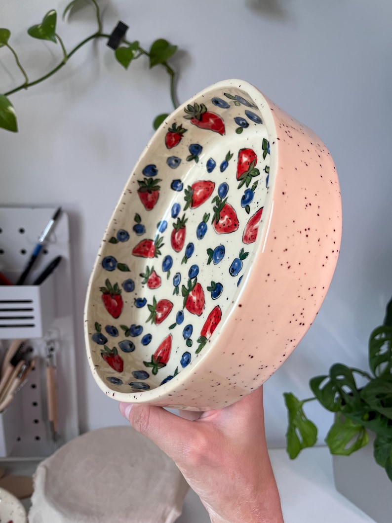 Op de afbeelding: Een ovale keramische kom met een lichtroze buitenkant, bespikkeld met zwarte stippen. De binnenkant heeft een witte achtergrond met een patroon van rode aardbeien en blauwe bosbessen. De kom wordt in een hand gehouden.