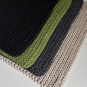Puede incluir: Primer plano de cuatro pa&ntilde;os de cocina de punto apilados. El pa&ntilde;o superior es negro con un borde verde, seguido de un pa&ntilde;o gris oscuro y un pa&ntilde;o beige. Los pa&ntilde;os de cocina est&aacute;n hechos de un hilo grueso y texturizado.