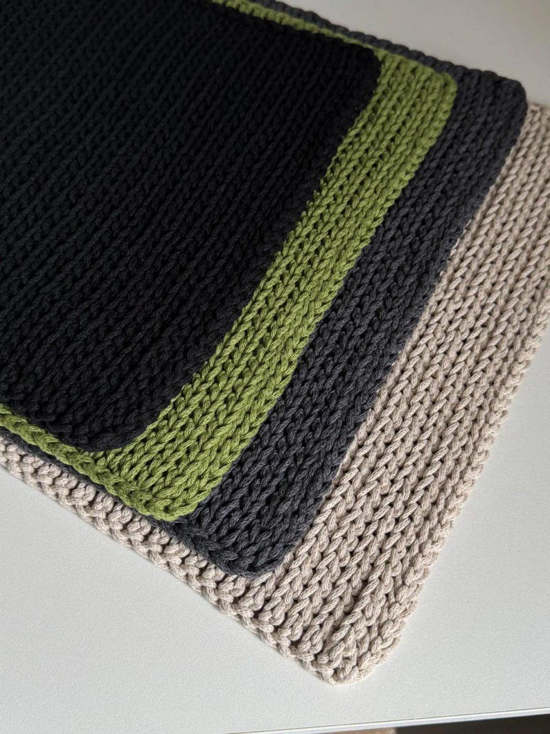 Puede incluir: Primer plano de cuatro pa&ntilde;os de cocina de punto apilados. El pa&ntilde;o superior es negro con un borde verde, seguido de un pa&ntilde;o gris oscuro y un pa&ntilde;o beige. Los pa&ntilde;os de cocina est&aacute;n hechos de un hilo grueso y texturizado.