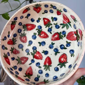 Puede incluir: Un tazón de cerámica blanca con un esmalte moteado presenta un diseño pintado a mano de fresas rojas y arándanos azules.